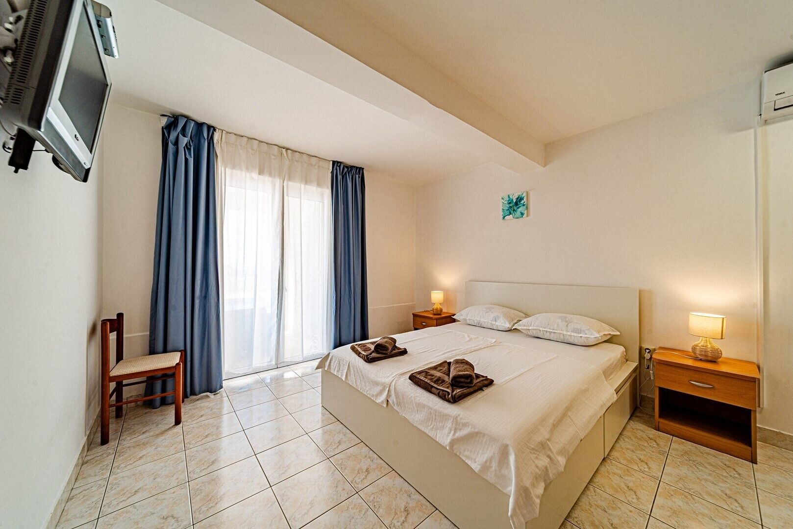 Une chambre d’hôtel comprenant un lit, deux tables de chevet, une chaise, un téléviseur fixé au mur et une fenêtre avec des rideaux.