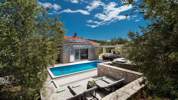Un espace aménagé au bord de la piscine, avec un mur en pierre, des chaises longues et un petit bâtiment doté d’une porte en verre.