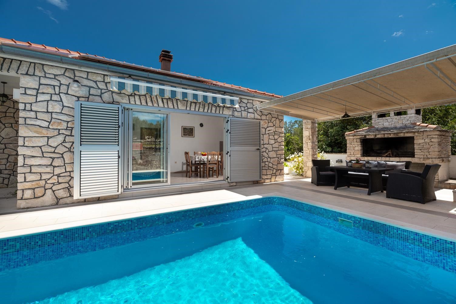 Une piscine avec un revêtement extérieur en pierre, un patio couvert et un barbecue.