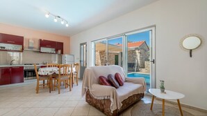 Un salon confortable avec un canapé, une table à manger et des chaises. On y trouve une petite table ronde agrémentée d’une plante. Une grande fenêtre offre une vue sur une piscine et un bâtiment en pierre.