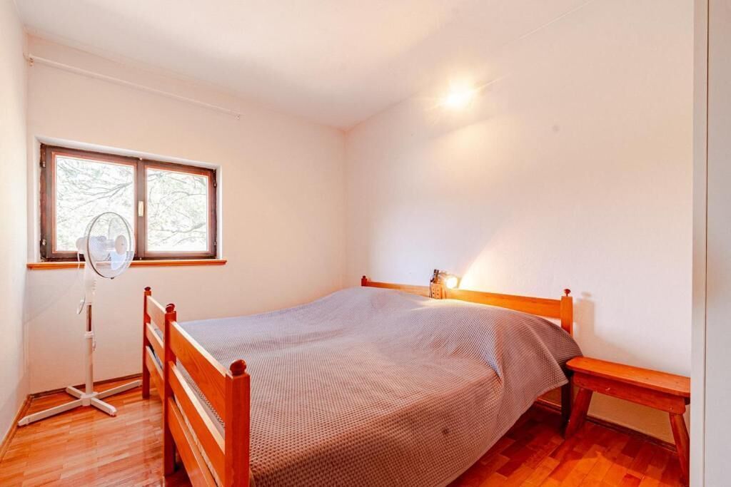 A bedroom with a wooden bed, a bedside table, a standing fan, and a window with a view of trees.