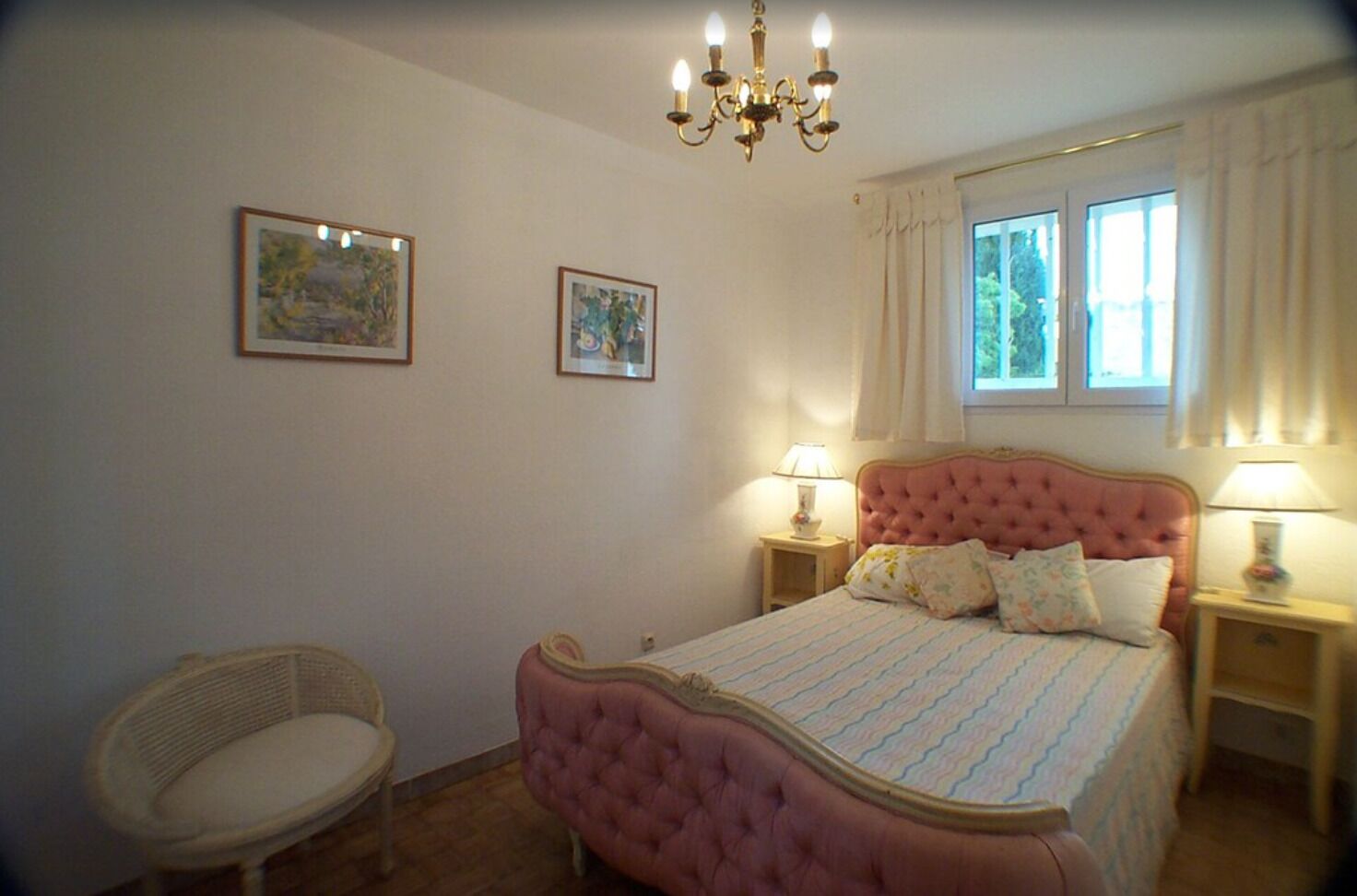 Una camera da letto con un letto trapuntato rosa, due comodini con lampade, una poltrona in vimini e due quadri appesi al muro.