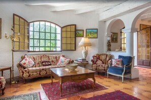 A traditional living room with a sofa, armchairs, a coffee table, and a lamp. There is a large window with a view of greenery outside. - Unique hilltop villa in Provence, stunning private pool, 6+bdrms + optional gite (Beaucaire)