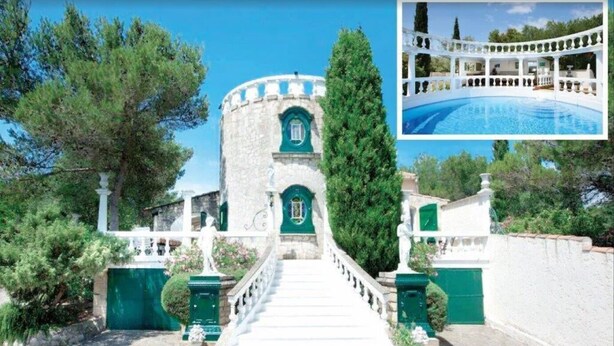 Un edificio in pietra con una torre centrale, una scalinata che conduce all'ingresso e una zona piscina recintata da una staccionata bianca.