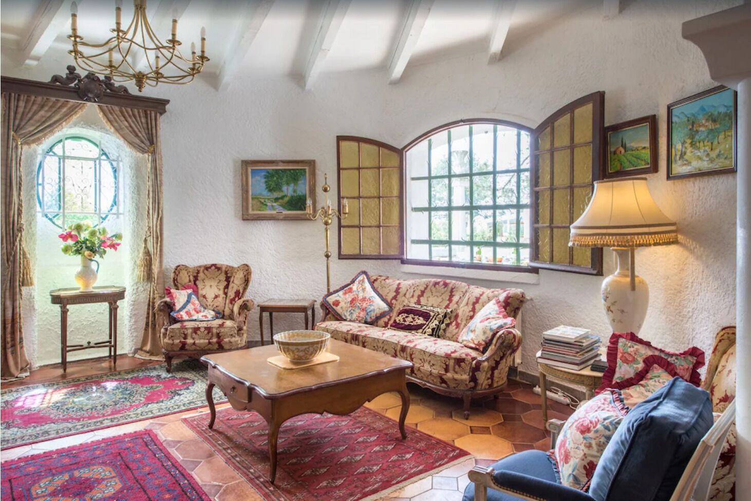 A cozy living room with a sofa, armchairs, a coffee table, and a lamp. There are windows with curtains, a chandelier, and a rug.