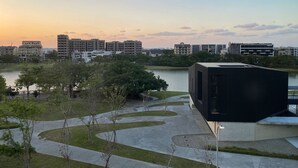 Um edifício moderno com telhado plano e fachada escura extensa, rodeado por área ajardinada com caminhos e árvores, com vista para um corpo de água e um horizonte urbano com diversos edifícios.