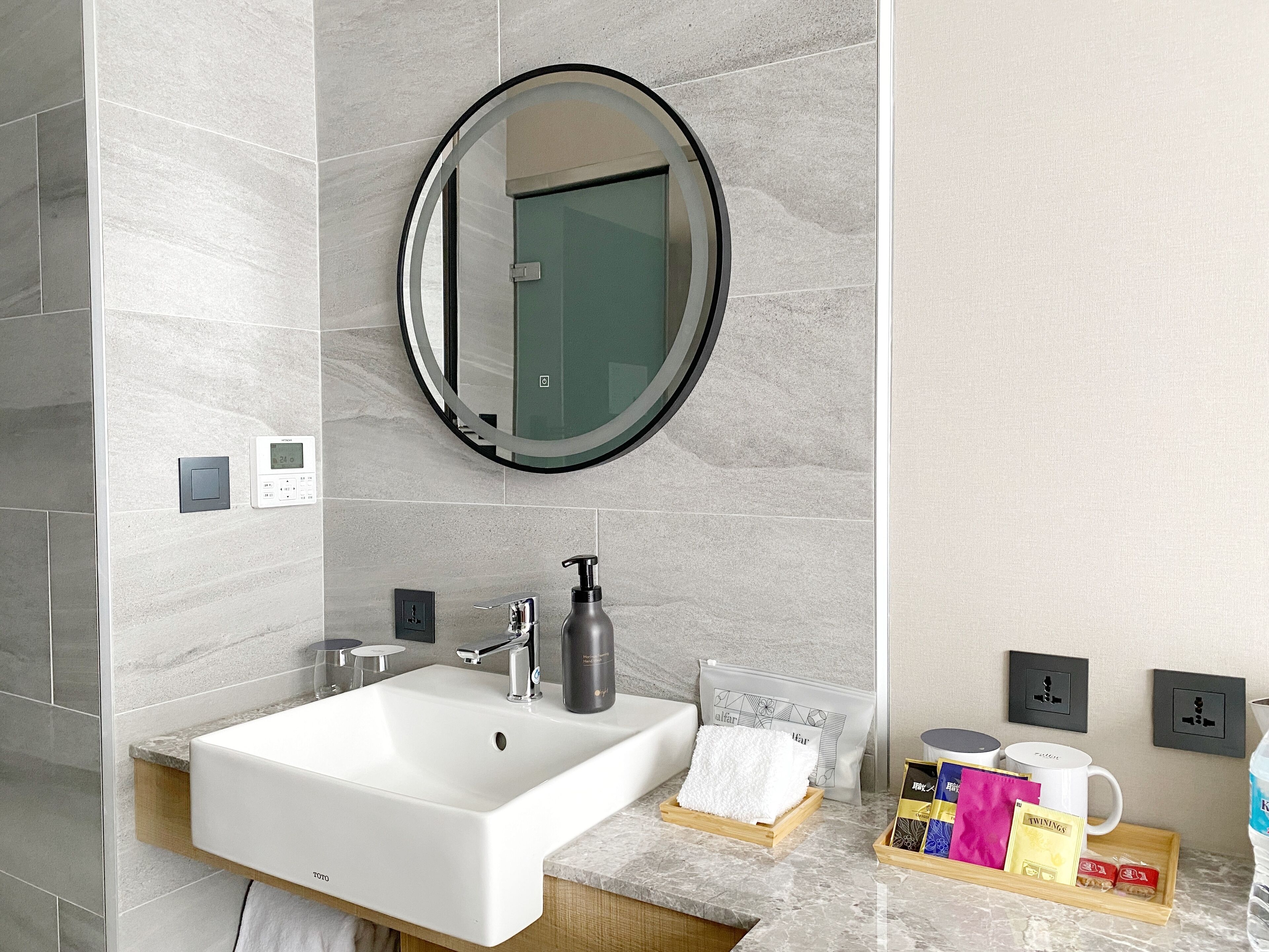 A modern bathroom with a white sink, a round mirror, a soap dispenser, and a towel rack. There are toiletries on the counter and a small tray with a towel and a bag.