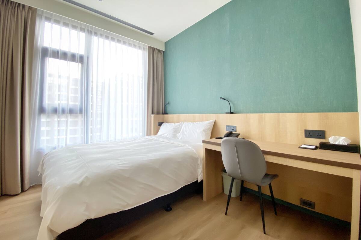 Une chambre d’hôtel avec un lit, un bureau et une chaise. Il y a une fenêtre avec des rideaux, un téléphone fixé au mur et une prise de courant.