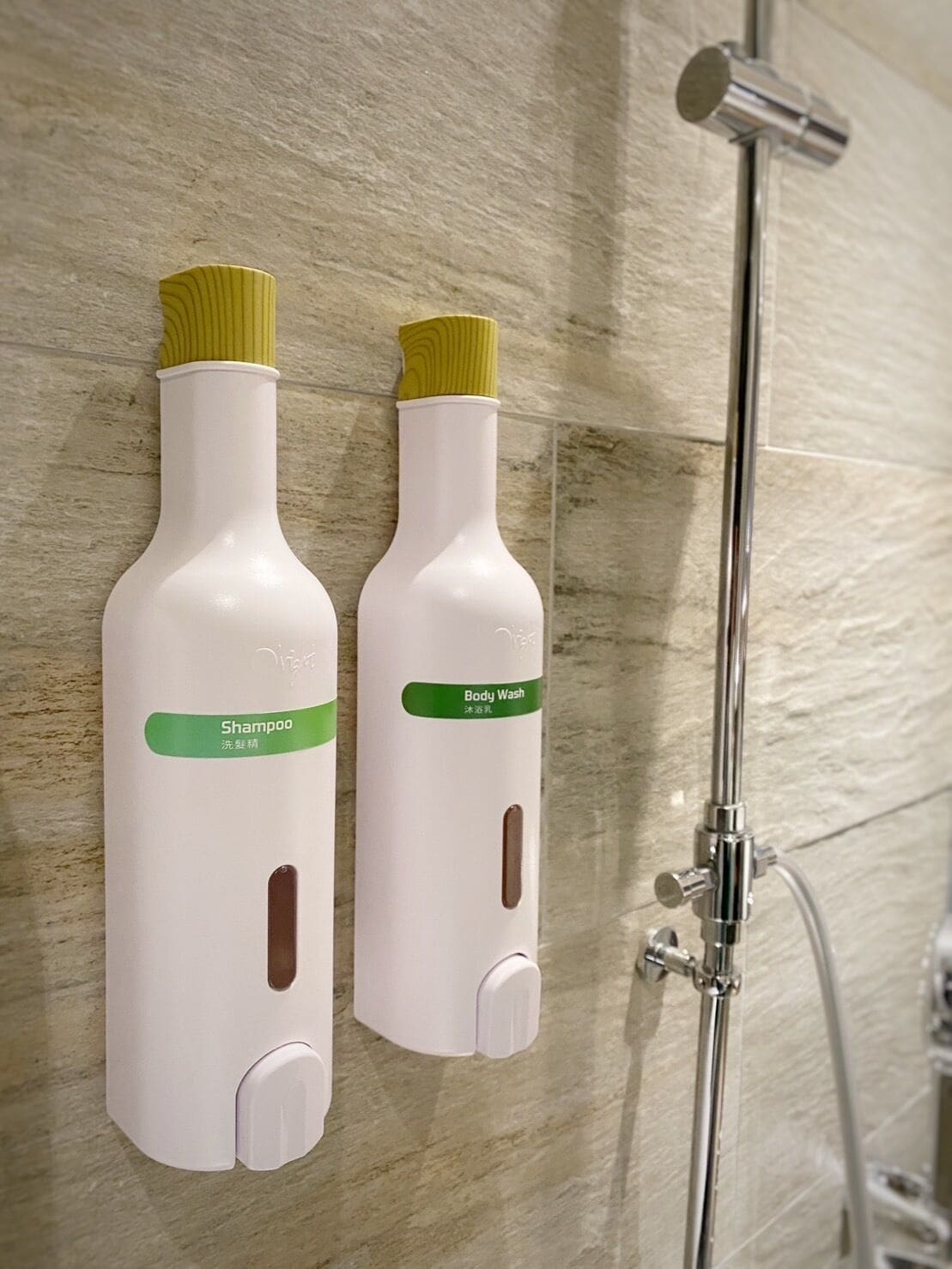 Two white bottles with yellow caps labeled "Shampoo" and "Body Wash" mounted on a wall next to a shower.