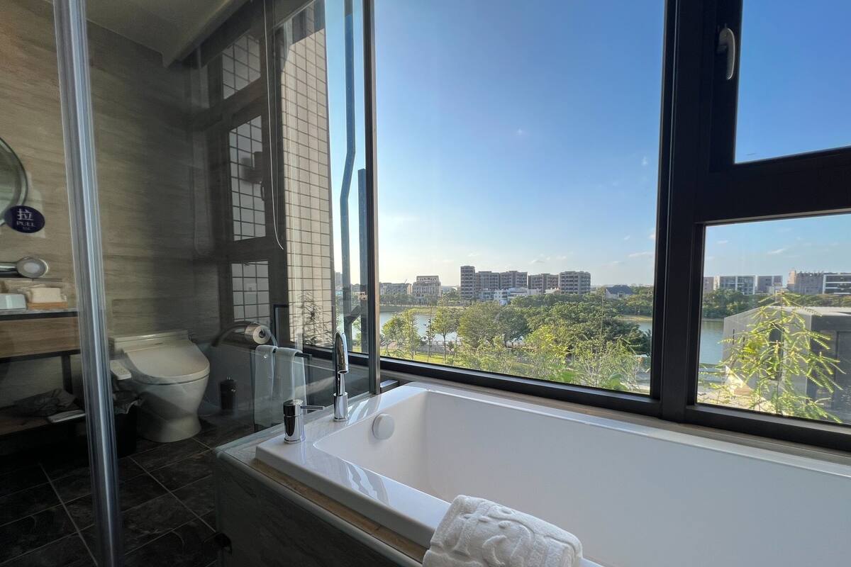 Une salle de bain moderne équipée d’une baignoire, de toilettes et d’un lavabo, et dotée d’une grande fenêtre offrant une vue sur la ville.