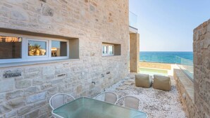 A stone building with a glass table and chairs, a view of the sea, and a balcony with cushions.
