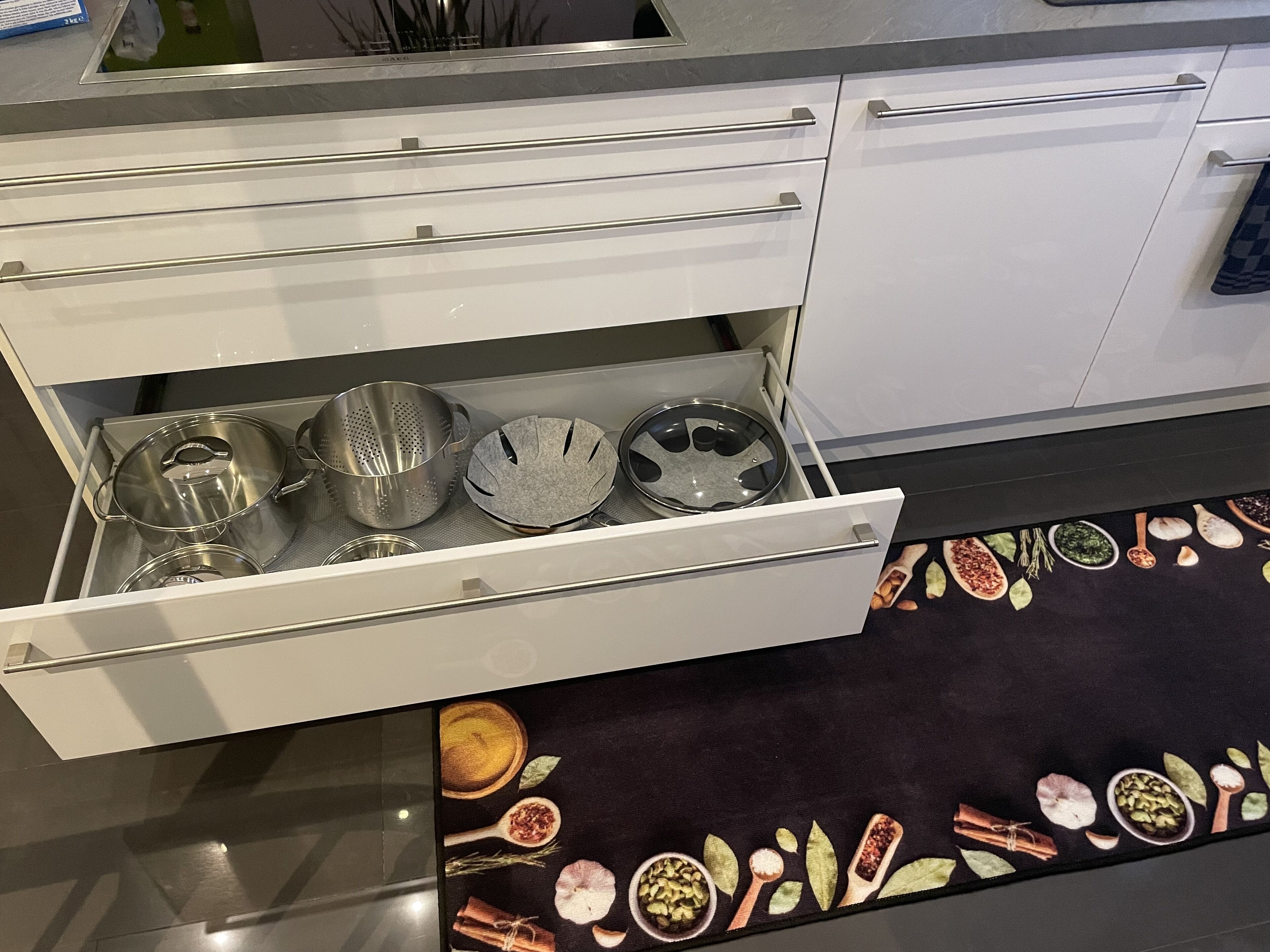 Una cucina con un cassetto pieno di utensili da cucina e un tappetino nero decorato con illustrazioni di cibo.