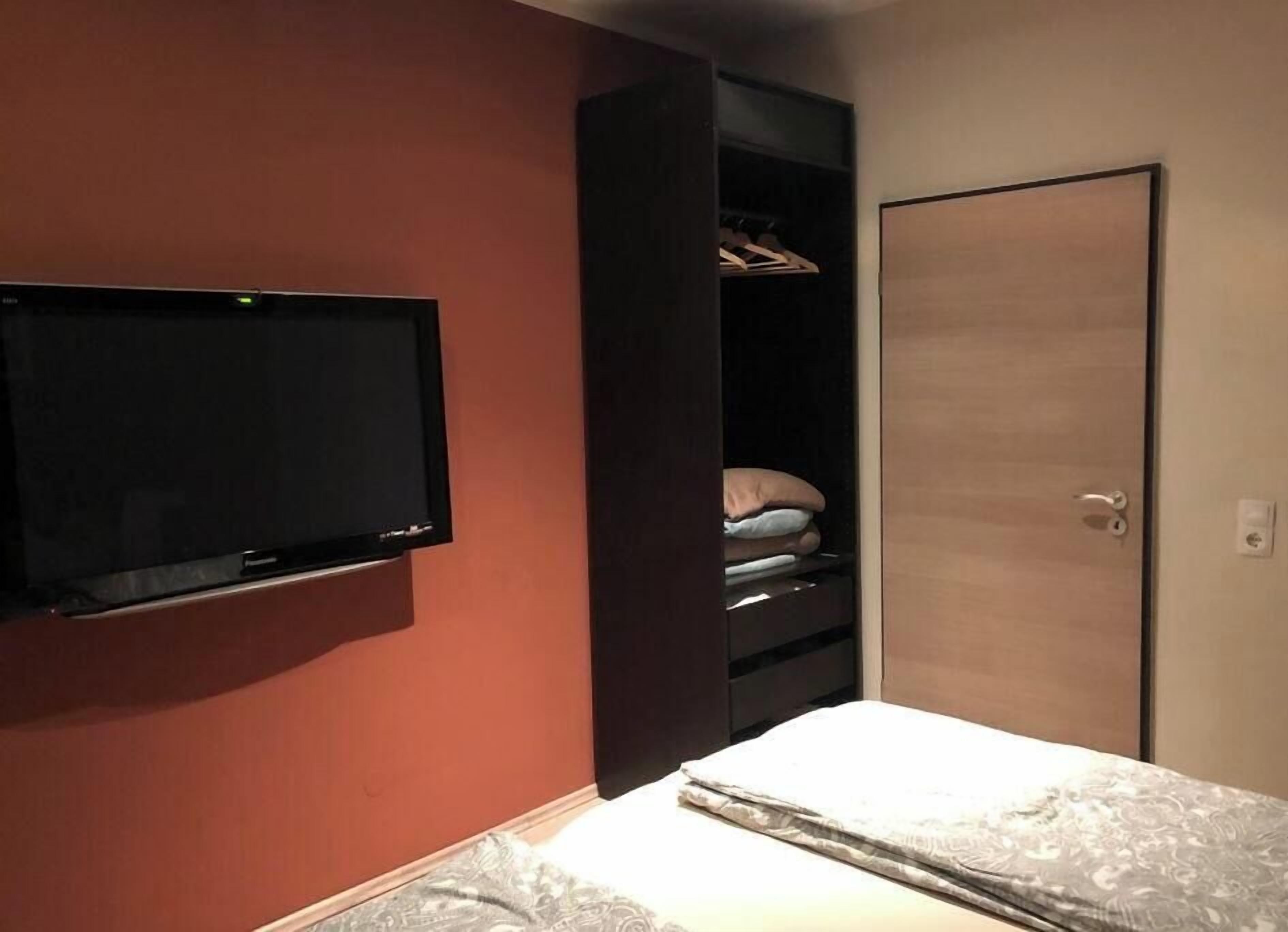 Ein Schlafzimmer mit an der Wand montiertem Fernseher, einem Kleiderschrank mit Kleidung und einer Tür.
