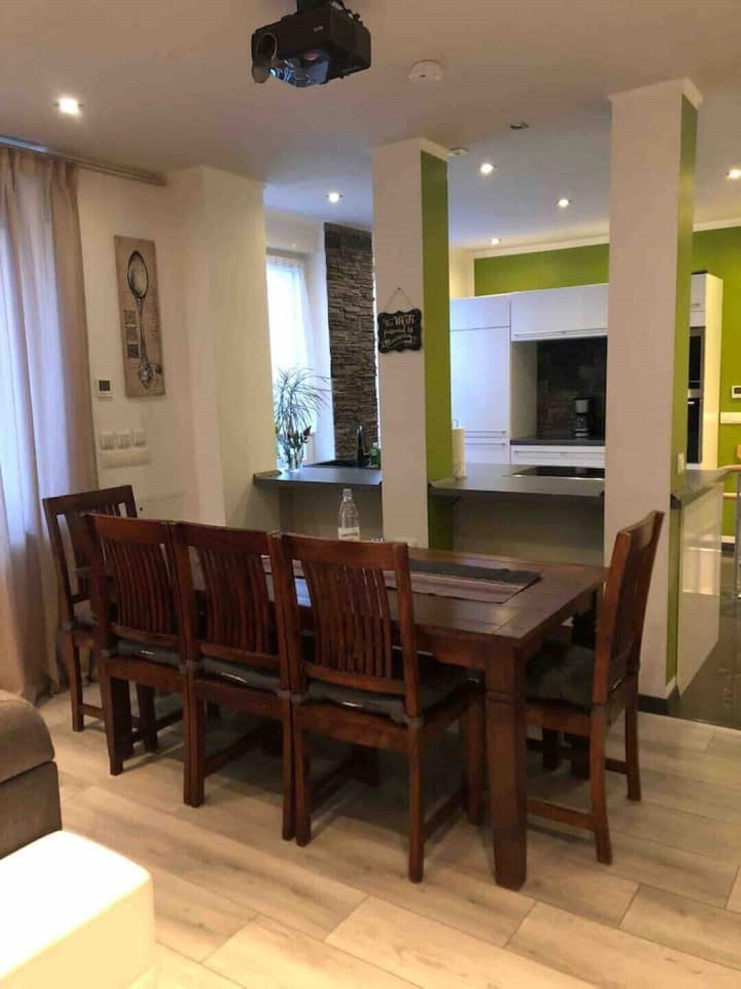 A dining area with a wooden table and chairs, a kitchen in the background, and a green accent wall.