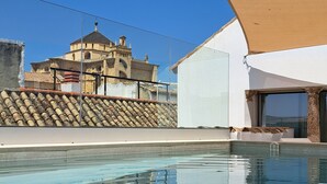 A swimming pool with a clear reflection, a large building with a tiled roof in the background, and a beige awning providing shade. - Casa del Cardenal (Córdoba)