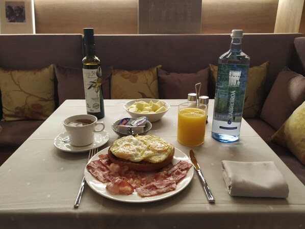 Une table dressée pour le petit-déjeuner avec une assiette d’œufs, de bacon et de pain grillé, une tasse de café, une bouteille d’huile d’olive, une bouteille d’eau et un bol de fruits, le tout sur une table dans une pièce chaleureuse, agrémentée d’un canapé et de coussins.
