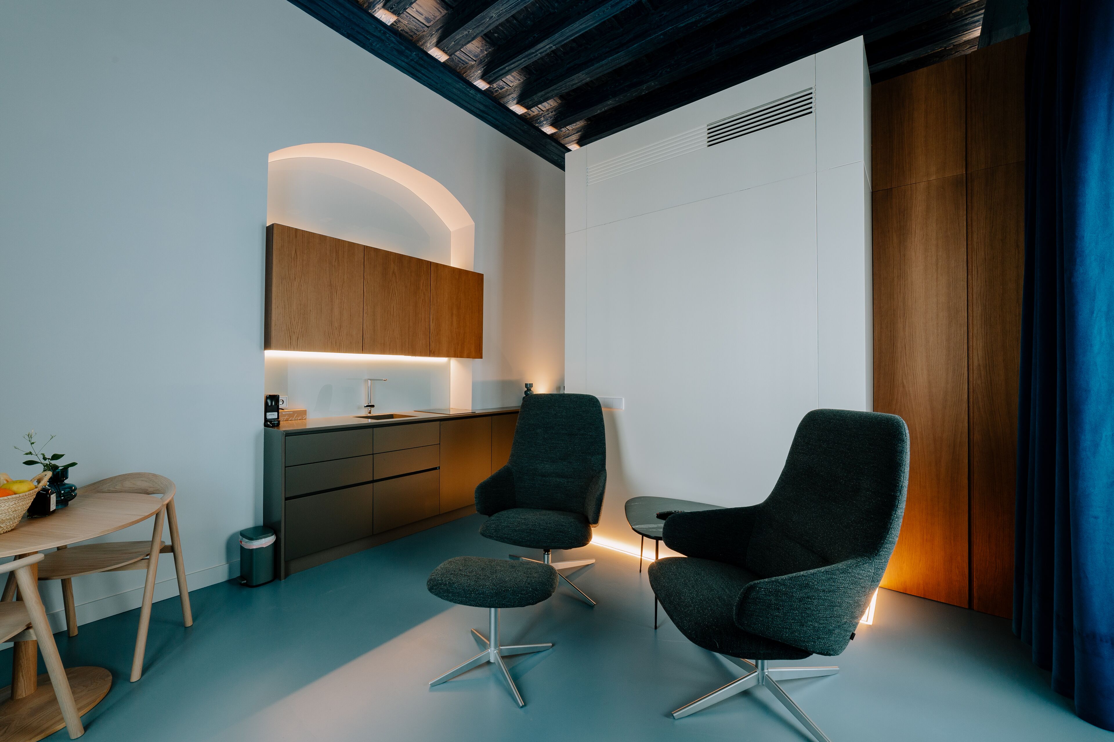 A modern, minimalistic room with a wooden cabinet, two dark armchairs, a small table, and a desk with a basket of fruit.