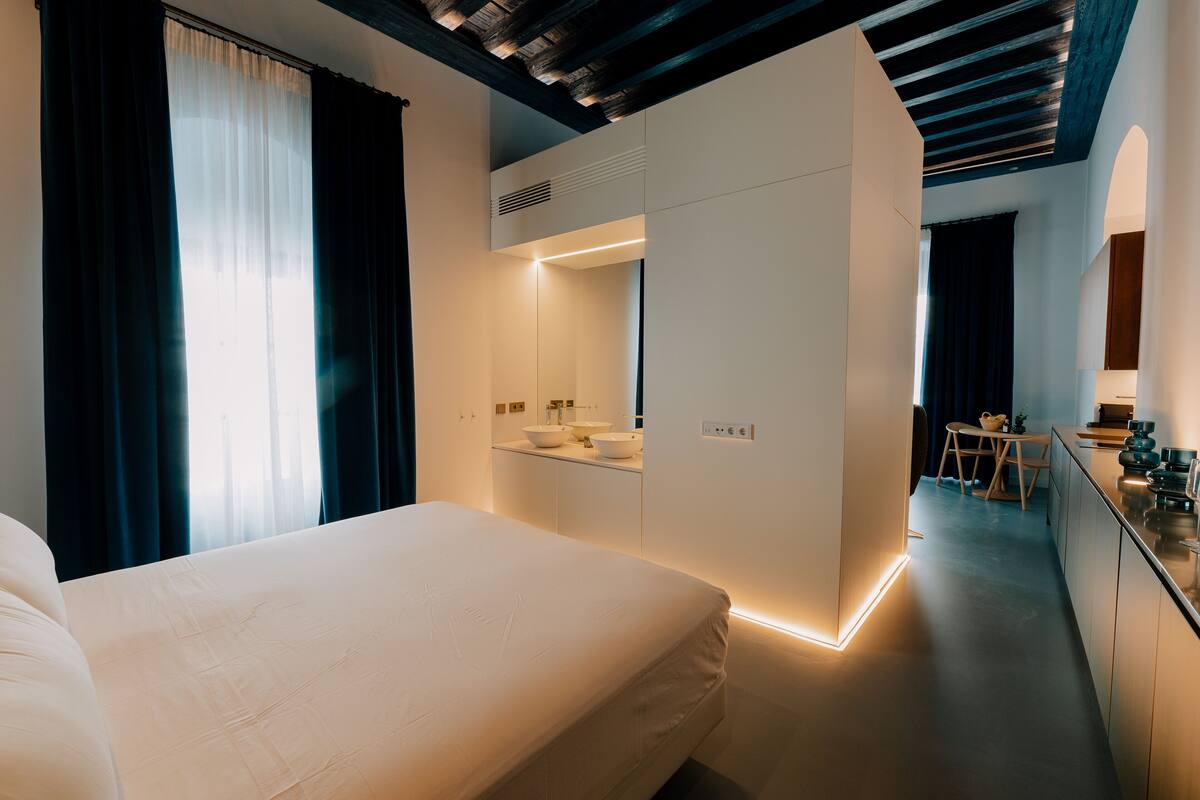 A modern hotel room with a large bed, a built-in wardrobe, a small kitchenette, and a dining area.