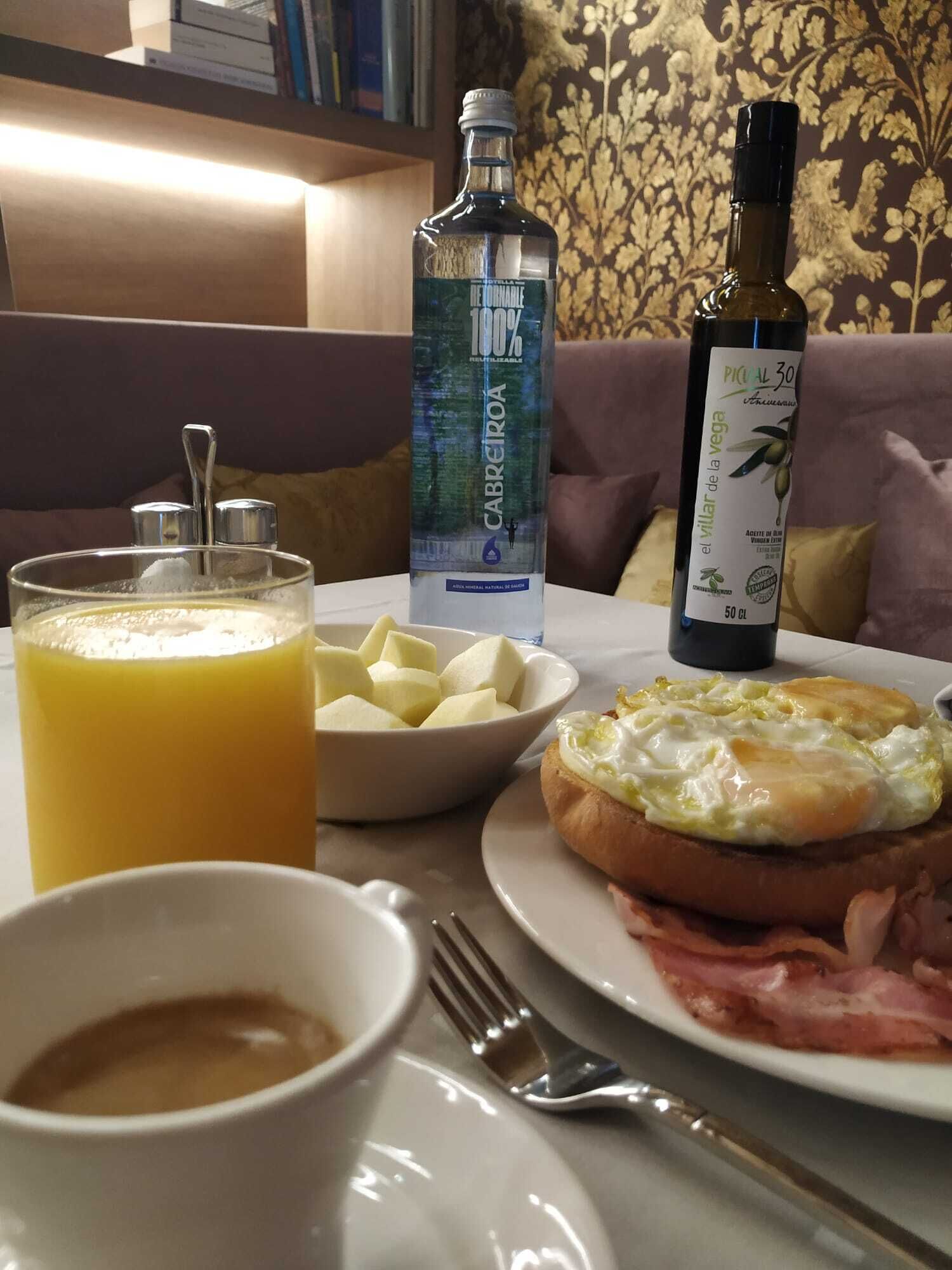 Une table mise pour le petit-déjeuner avec un verre de jus d’orange, un bol de fromage, une assiette avec des œufs et du jambon, une bouteille d’huile d’olive et une bouteille d’eau.