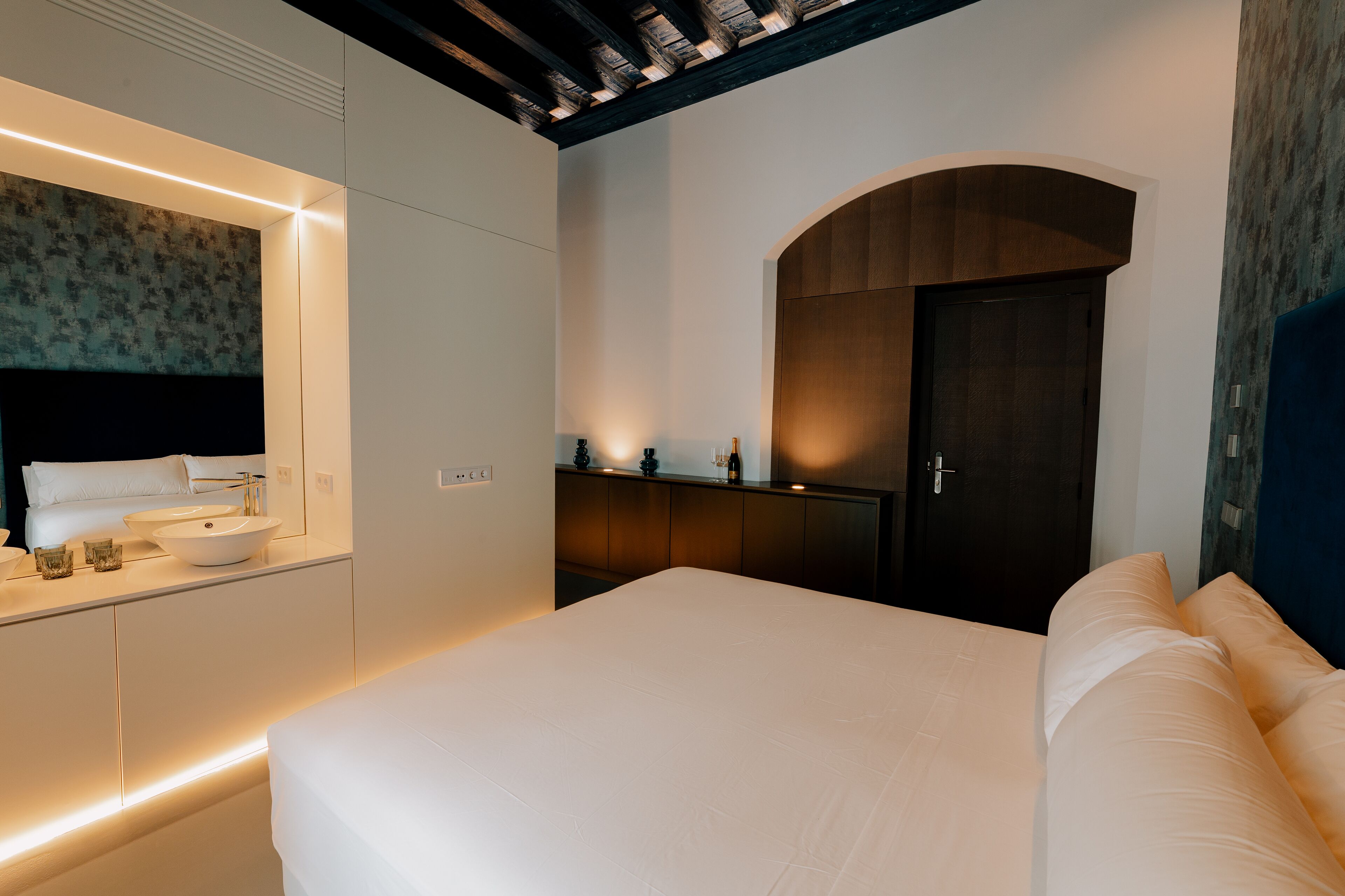 Un dormitorio moderno con una cama grande, una mesita de noche, un armario empotrado y un baño con un espejo grande y un lavabo.
