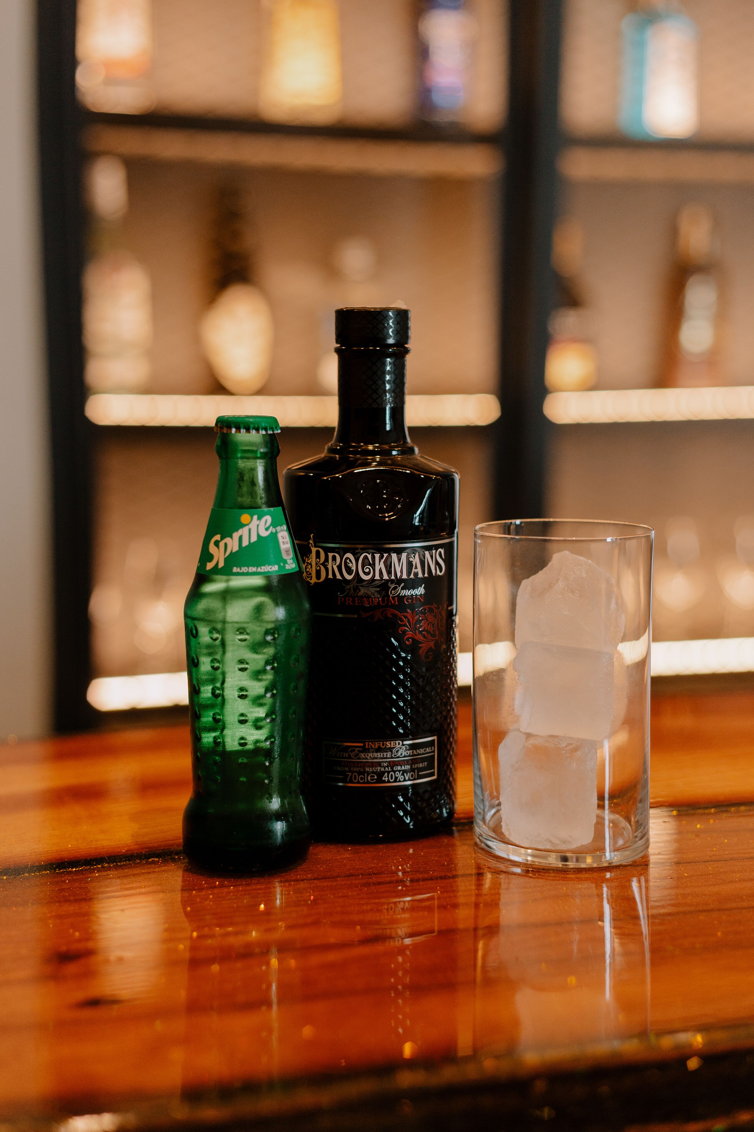 Una botella de Sprite, una botella de Bockmanns y un vaso con hielo sobre una superficie de madera.