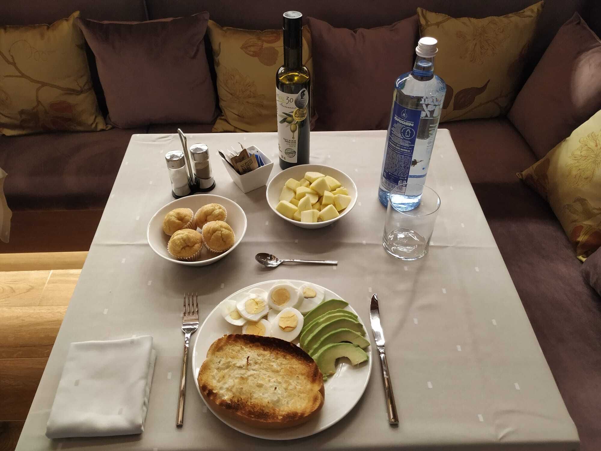 A dining table with a meal consisting of bread, eggs, avocado, and potatoes, along with a bottle of water and a bottle of wine.
