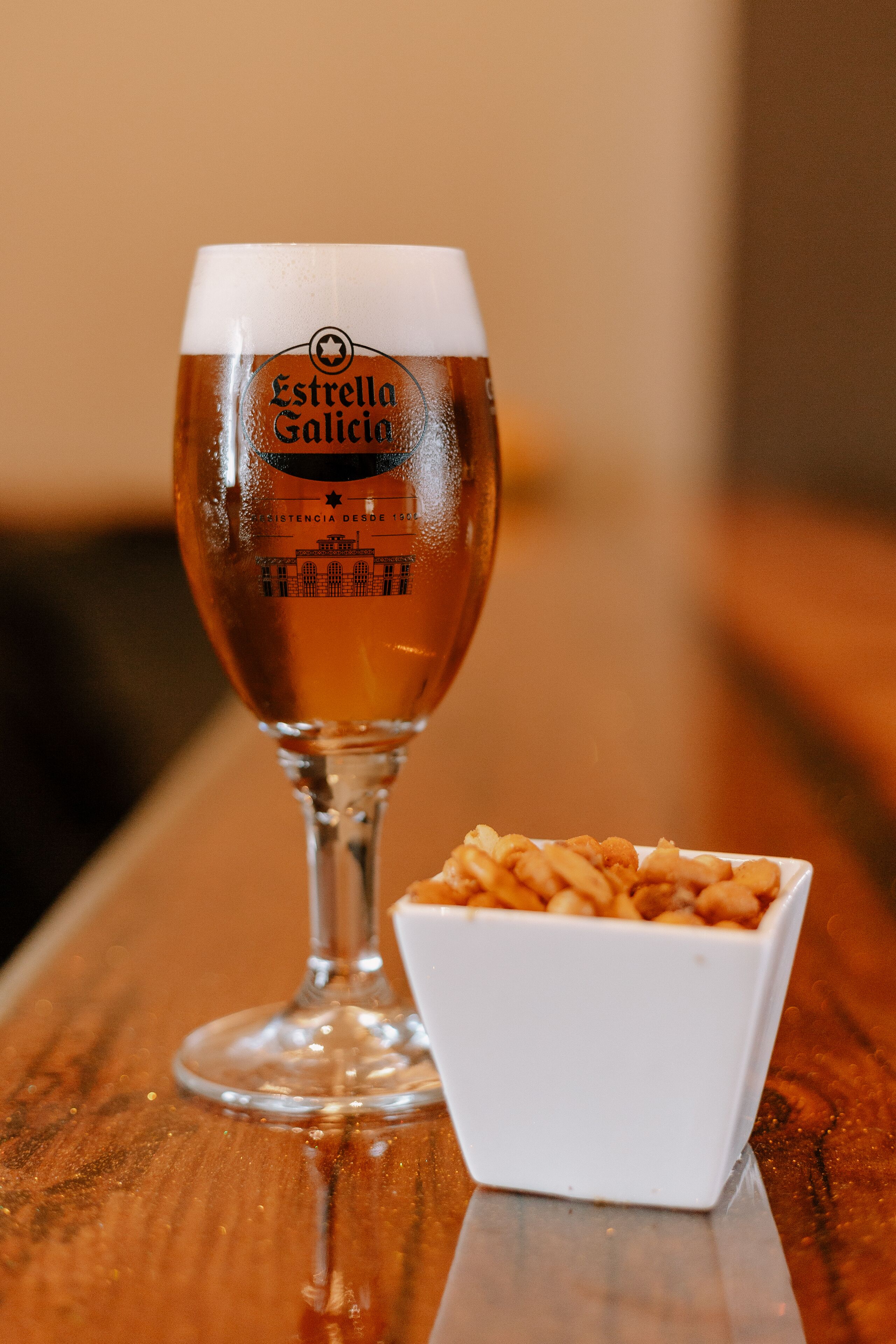Una cerveza con un logotipo y un tazón pequeño de aperitivos sobre una superficie de madera.
