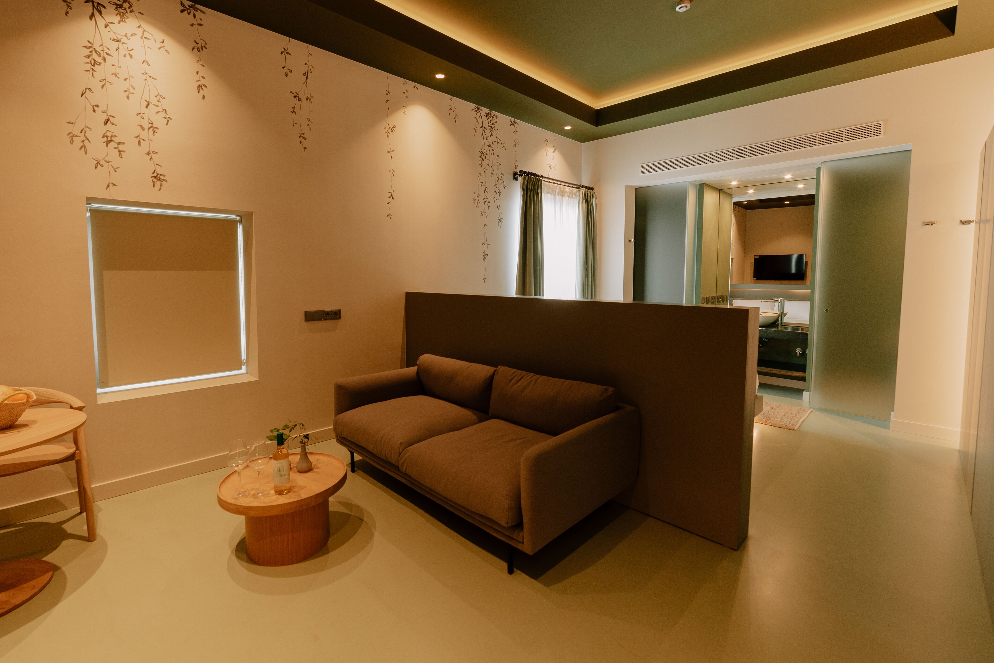 Una sala moderna con un sofá marrón, una mesa de centro de madera y una planta pequeña. Hay una ventana con vistas al exterior y un televisor montado en la pared.