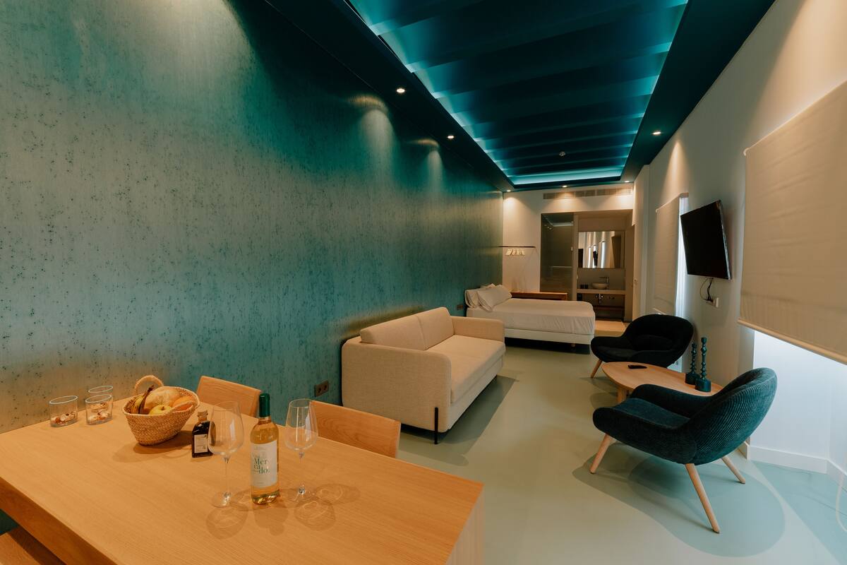 A modern living room with a wooden dining table, a sofa, a dark armchair, and a flat-screen TV mounted on the wall.