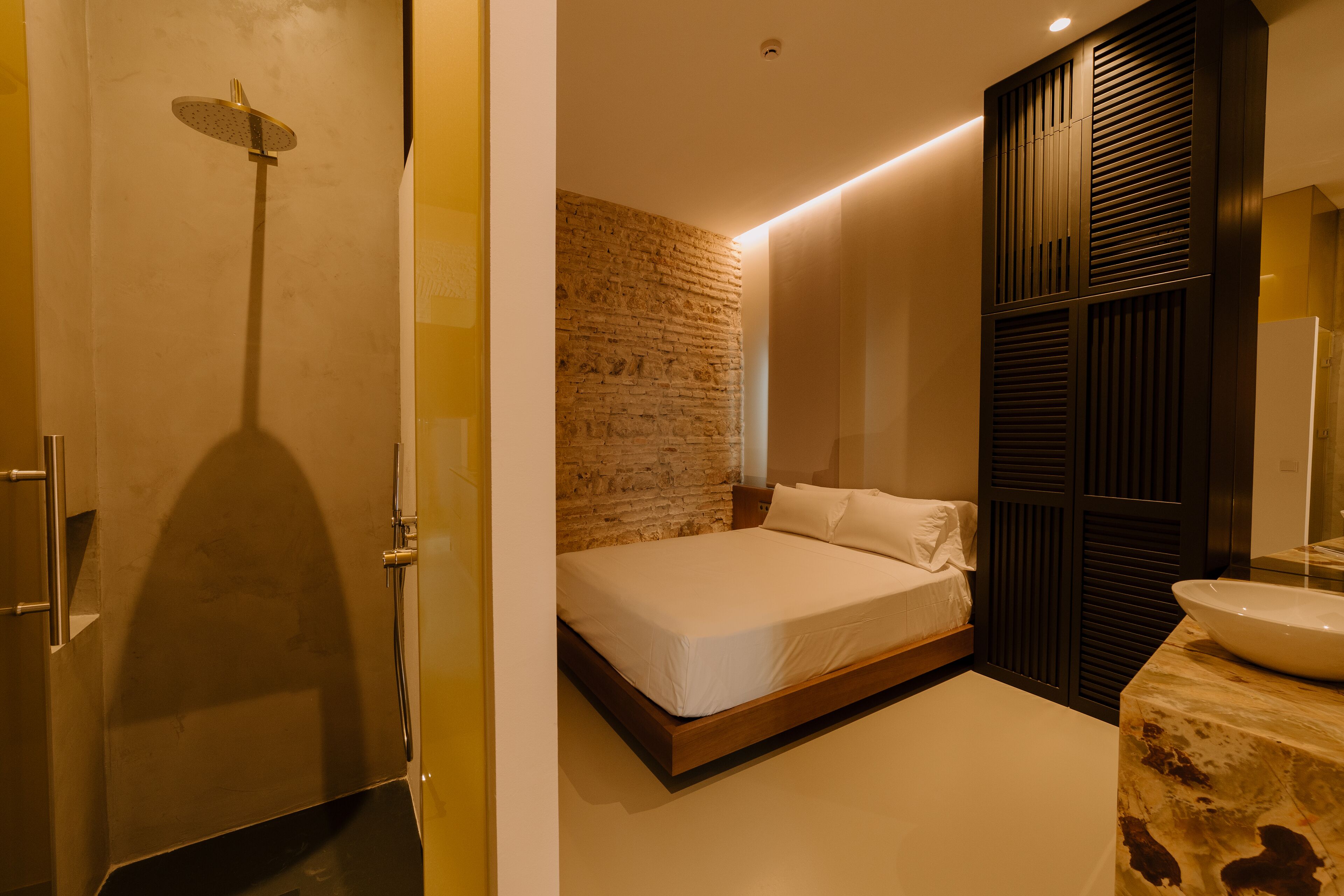 Une chambre moderne avec un mur en briques, un lit, une tête de lit en bois, une salle de bain avec un lavabo et un miroir, et une douche.