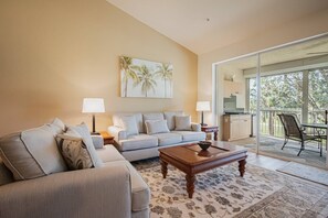 A living room with a sofa, a coffee table, and a painting of palm trees. There is a balcony with a view of trees.