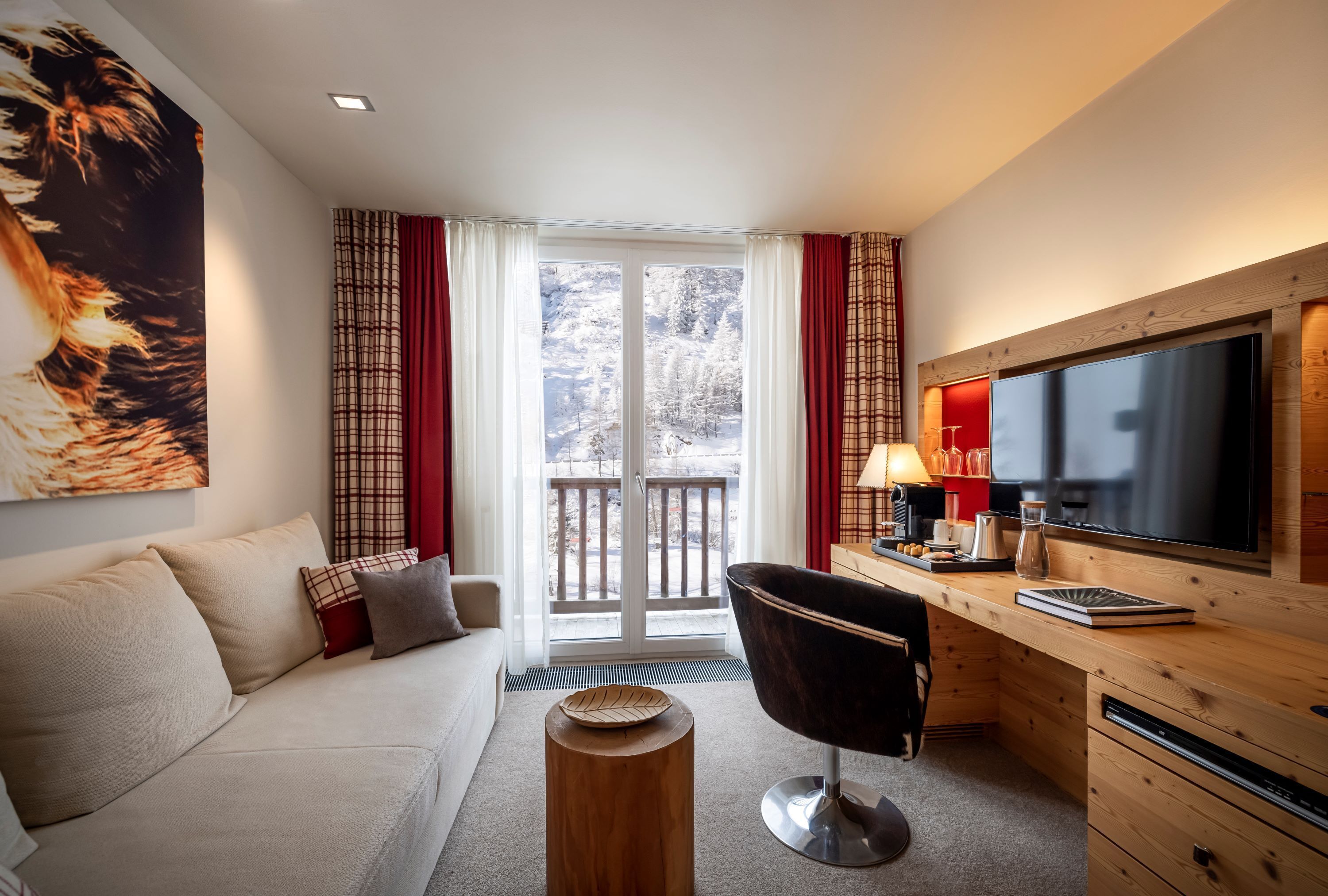 A modern hotel room with a sofa, a desk with a flat-screen TV, a chair, and a large window with a view of a snowy landscape.