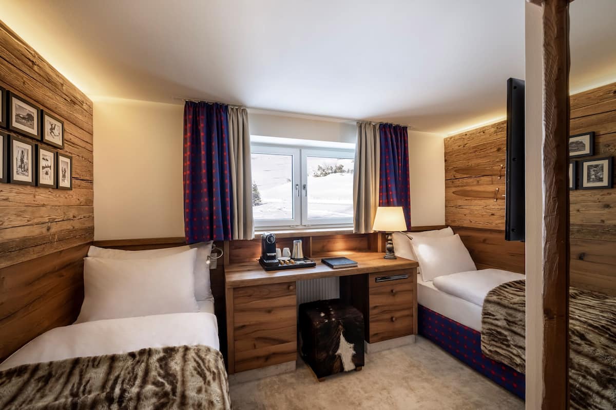 A cozy room with wooden walls, a desk with a lamp, two beds, and a window with curtains.