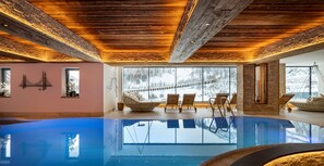 An indoor swimming pool with wooden ceiling beams, large windows with a snowy view, and lounge chairs. - Kesselspitze Hotel & Chalet, Valamar Collection (Tweng)
