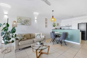 Un salon moderne avec une table basse en verre, un canapé avec des coussins, un coin repas avec des chaises et une cuisine équipée de placards et d’un réfrigérateur.