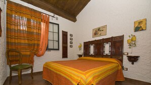 Une chambre avec un grand lit, une chaise en bois, une fenêtre avec des rideaux, une étagère fixée au mur avec un vase, et un tableau.