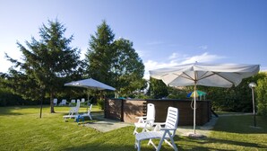 Un espace extérieur verdoyant avec des chaises longues blanches et des parasols, une structure en bois et des arbres en arrière-plan.