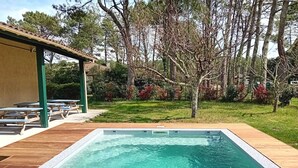 Une piscine rectangulaire entourée d’une terrasse en bois, un espace de détente couvert avec des bancs, et une pelouse avec des arbres et des arbustes.