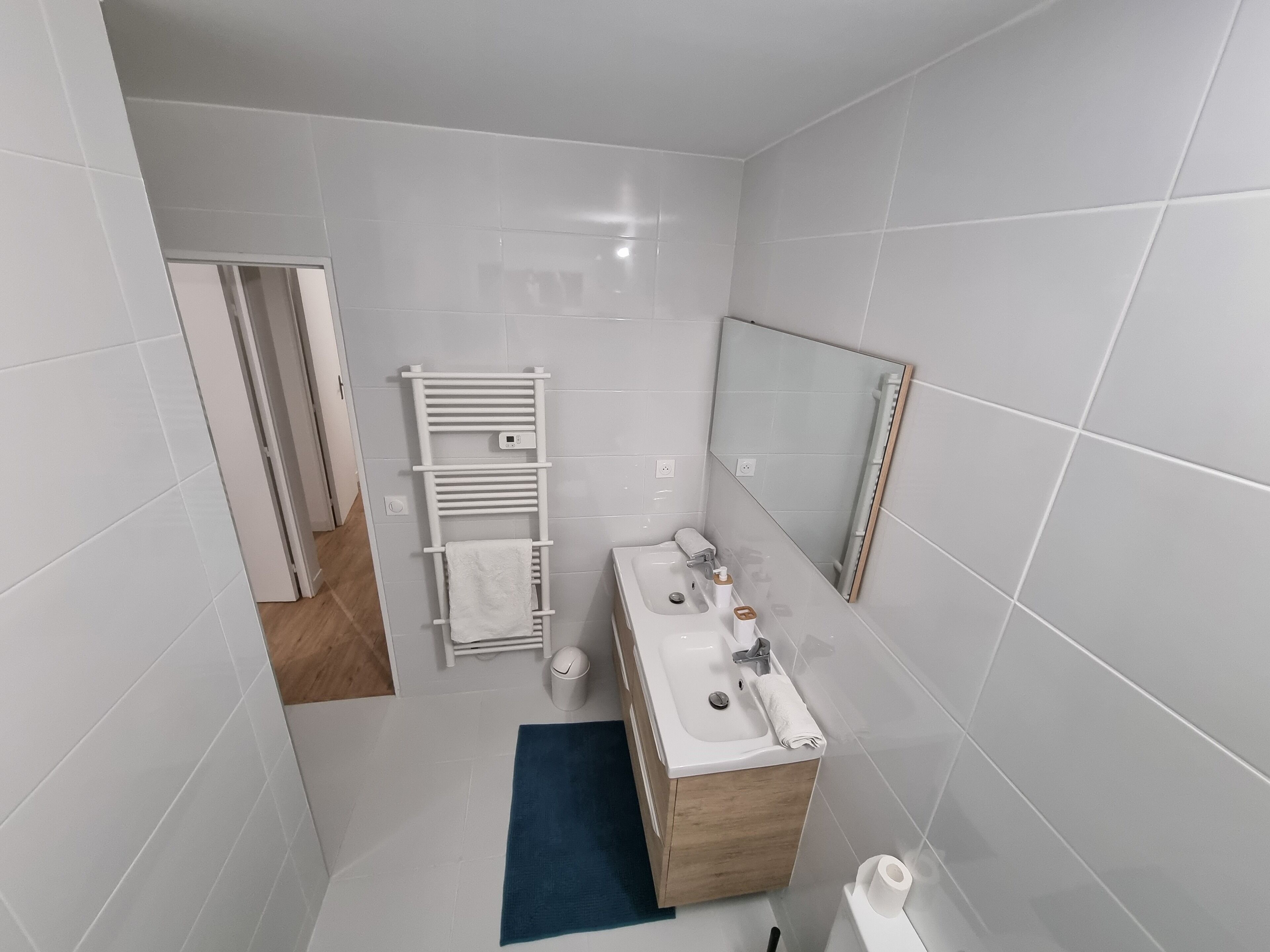 A modern bathroom with white tiles, a wooden vanity, a white sink, a mirror, and a towel rack.