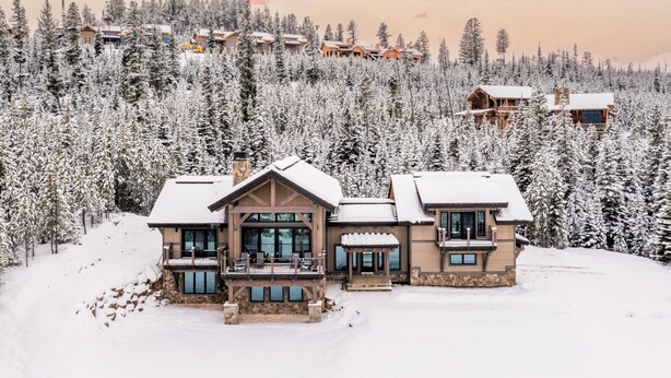A large, multi-story lodge with a stone facade and multiple balconies, surrounded by a dense forest of snow-covered trees.
