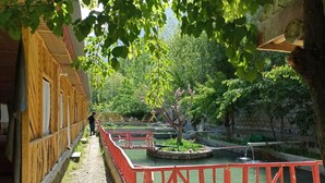 A narrow pathway flanked by wooden buildings, leading to a pond with a red railing.
