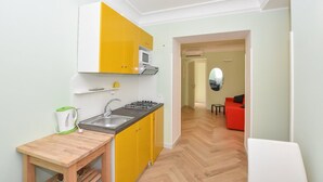 A compact kitchen with yellow cabinets, a microwave, and a gas stove. There is a small wooden table with a kettle and a sink. A red sofa and a mirror are visible in the adjacent living area. - Apartament CARPEDIEM in the Historical center, accessible by 59 steps At 650mt from the Cathedral (Amalfi)