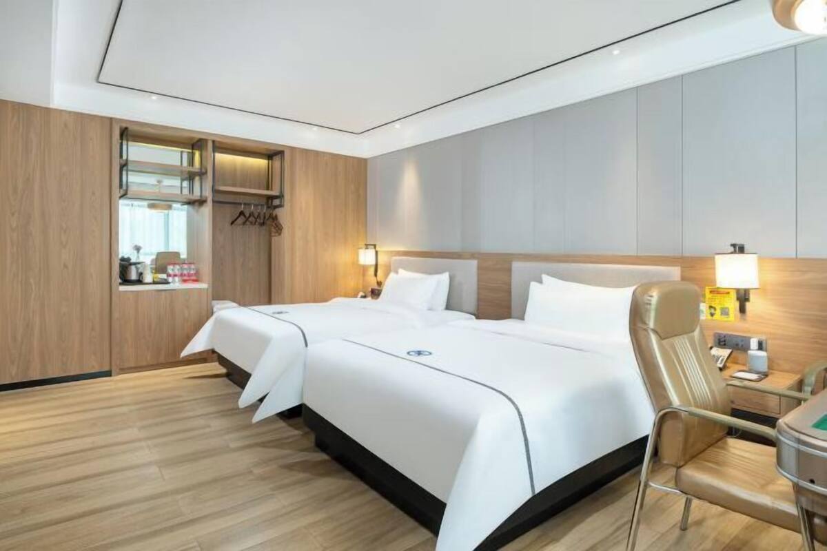 A hotel room with two beds, a wooden wardrobe, a small kitchenette, and a bedside table with a lamp.