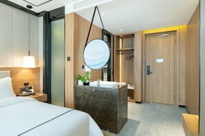 A modern hotel room with a bed, a marble bathroom vanity, a round mirror, and a wooden door.
