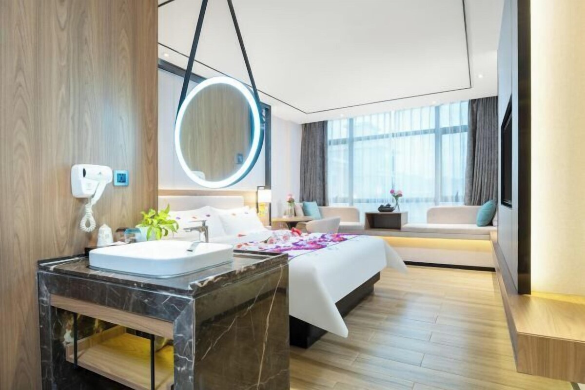 A modern hotel room with a marble bathroom, a large bed with a white bedspread, a round mirror, and a seating area with a view of the city.