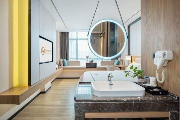 A modern hotel room with a marble bathroom vanity, a round mirror, a bed with a view, and a bedside table with a lamp and a hairdryer.