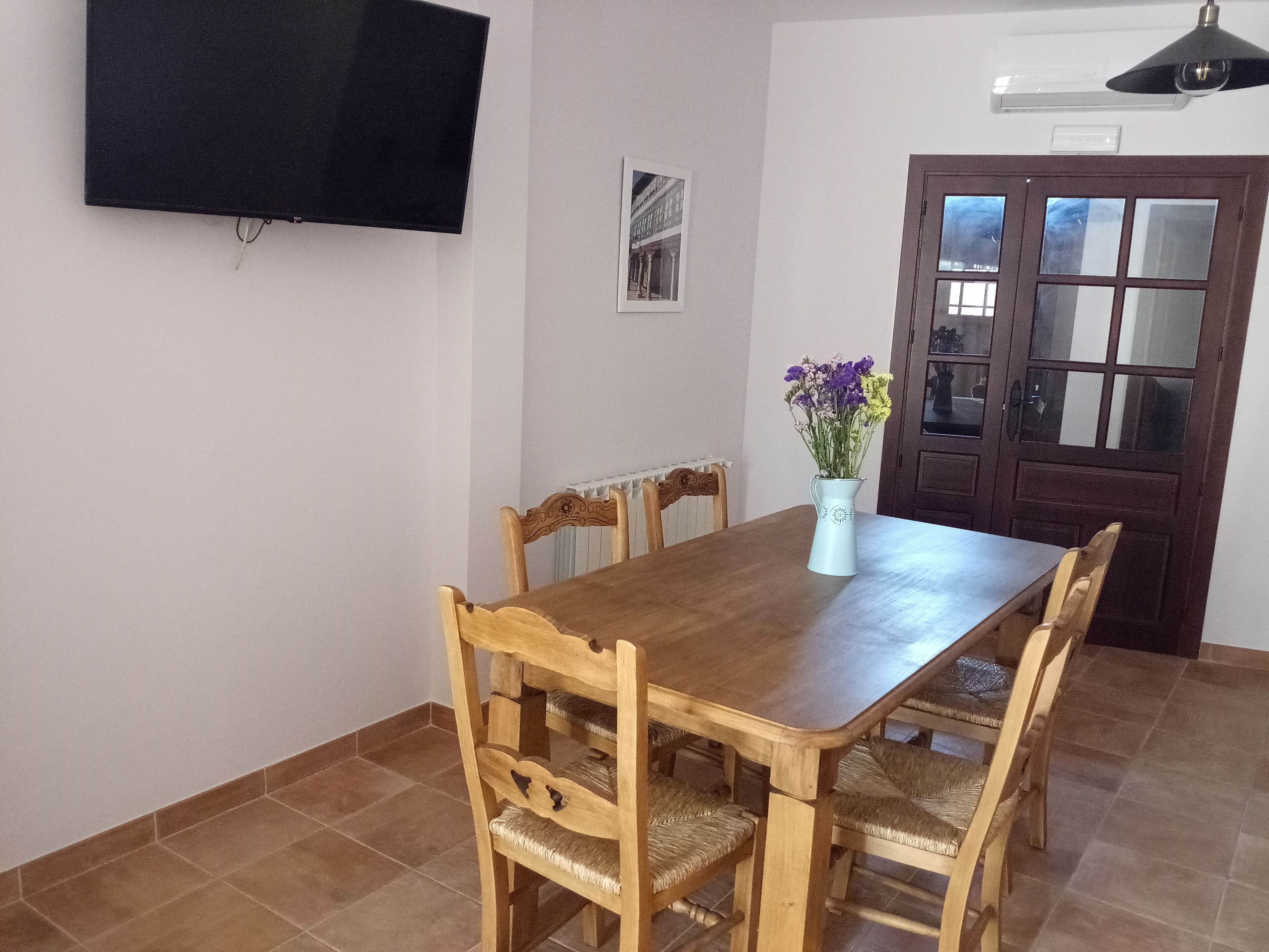 A dining area with a wooden table and chairs, a vase with flowers, a wall-mounted TV, and a door with glass panels.