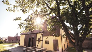 Un bâtiment avec un toit brun et des murs jaunes, un espace avec des bancs en bois et un arbre au premier plan.