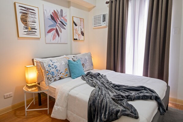 A neatly made bed with a white comforter and decorative pillows, a bedside lamp, and a gray throw blanket. - Delightful Apartment Holiday Rental (Davao City)