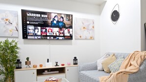 A modern living room with a flat-screen TV displaying a show, a sofa, a coffee table with books, a side table with decorative items, and a potted plant. - Delightful Apartment Holiday Rental (Davao City)